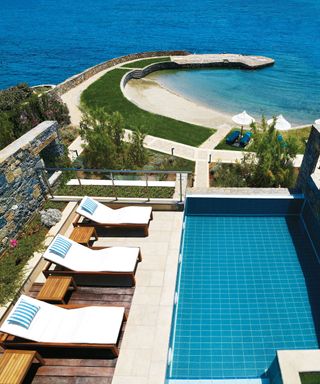 An example of pool patio ideas showing a rooftop swimming pool with a cream patio and a decking area overlooking the sea