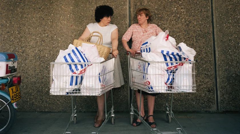 Two women stand side-by-side against a pebbled wall, each leaning on a shopping cart overflowing with white and blue plastic grocery bags.