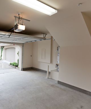 A white empty garage interior with white walls, two LED lights on the ceiling, gray skirting, a gray floor, and an open garage door opening up to show a white house