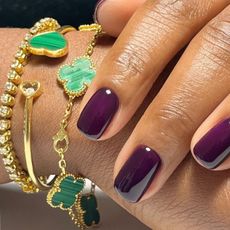 A woman's hands wearing green and gold jewellery with an aubergine manicure
