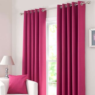 Fuchsia Solar Blackout Eyelet Curtains in a living room behind a white sofa which has a cushion on it in the same fuschia colour as the curtains