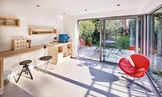 An old garage converted into a home office, with a wooden desk and shelving unit, a red desk chair and bifold doors, one of the best garage conversion ideas