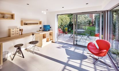 An old garage converted into a home office, with a wooden desk and shelving unit, a red desk chair and bifold doors, one of the best garage conversion ideas