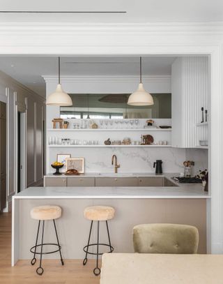 A small kitchen layout with white marble countertops and backsplash. The cabinets are painted in a beige color, the bar stools are beige and the pendant lights are beige.