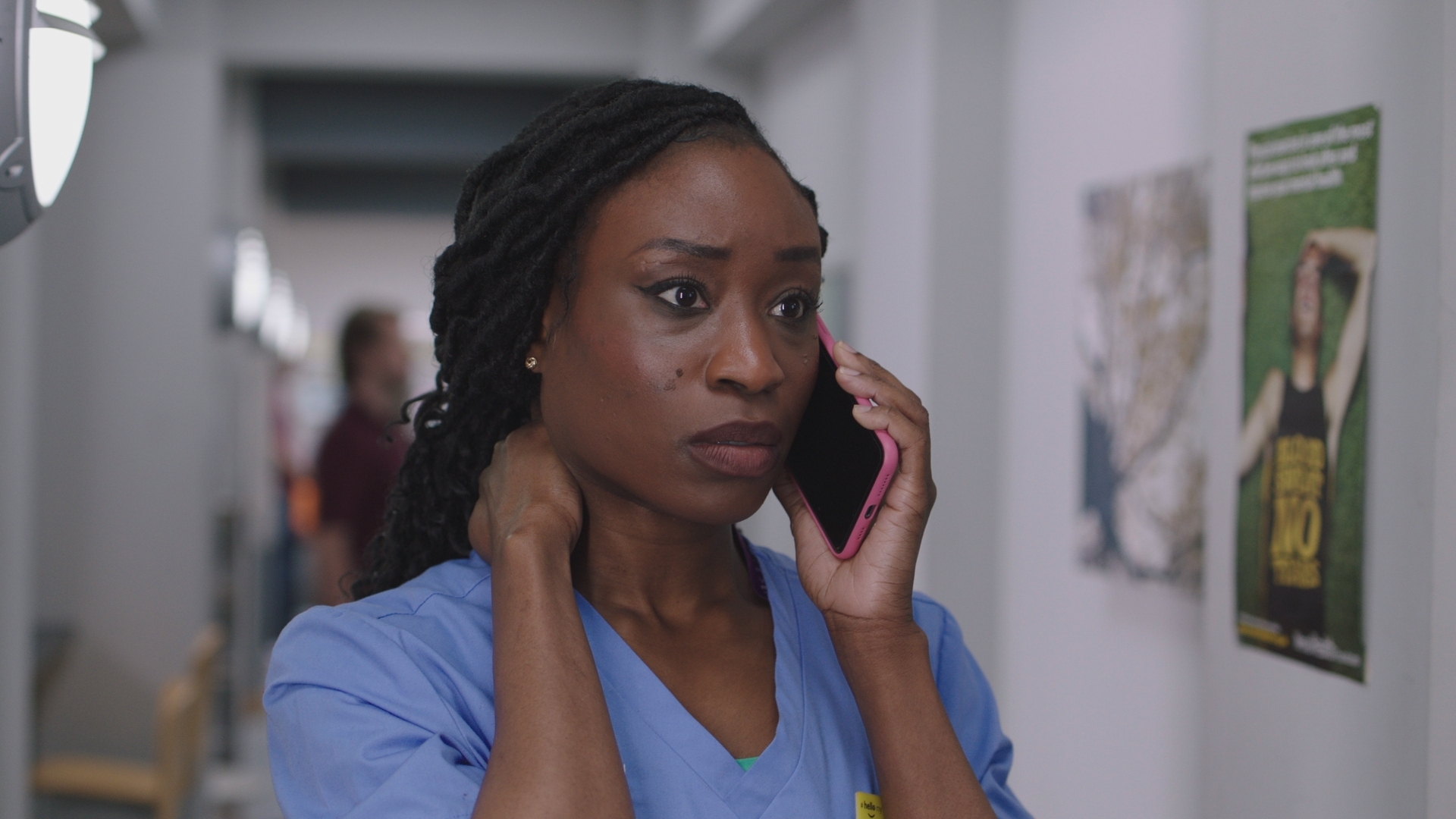 Ngozi at work, wearing blue scrubs, and on the phone looking troubled.