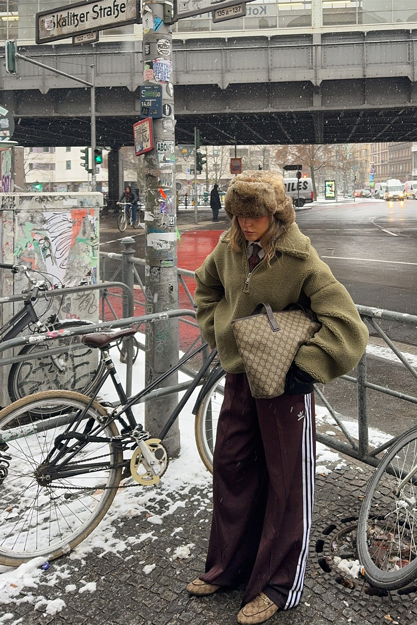Berlin fashion trends are shown in a street style photo of a woman standing outside near a bike rack wearing a fur hat with a green shearling jacket layered over a white button-down and red menswear tie, styled with brown track pants, Gucci loafers, and a Gucci tote bag