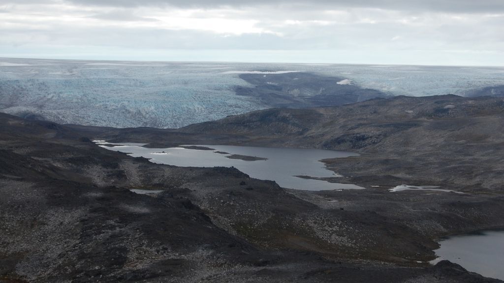 a landscape photo of an outcrop of Greenland&#039;s Isua supracrustal belt, shows valley with a pool of water in the center and a coastline and ocean beyond