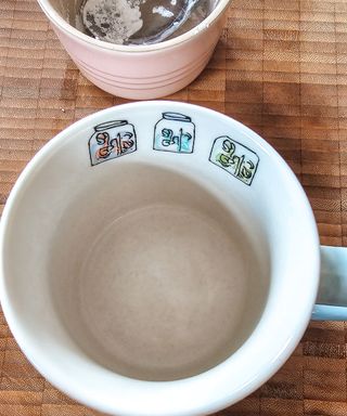 A birds eye shot of a white mug with a pink ceramic pot above it, all on a wooden chopping board