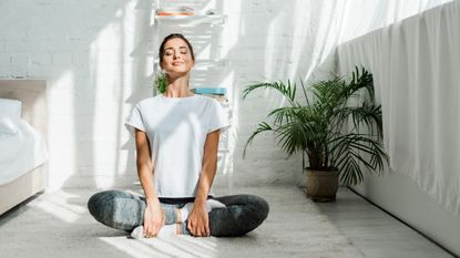 Woman in a cross legged position on the floor smiling as morning sunlight floods her stylish bedroom