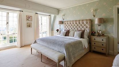 A mint green bedroom with a large bed with padded headboard and a green and white gingham bench at the end of it. To either side of the bed is a white chest of drawers, with matching silver lamps on top. To the left are two large floor-to-ceiling windows with open cream curtains open.