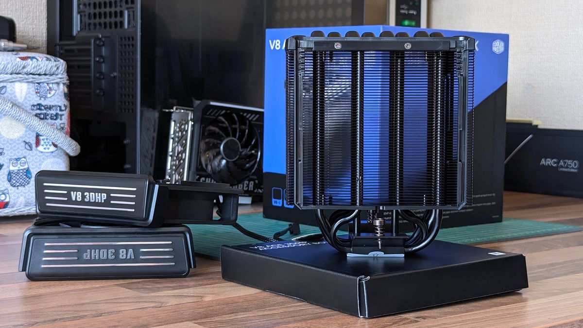 The Cooler Master V8 ACE 3DHP CPU cooler stands upright on a desk, displaying its heat pipes. Graphics cards, product boxes, and other hardware fill the background of the workspace.