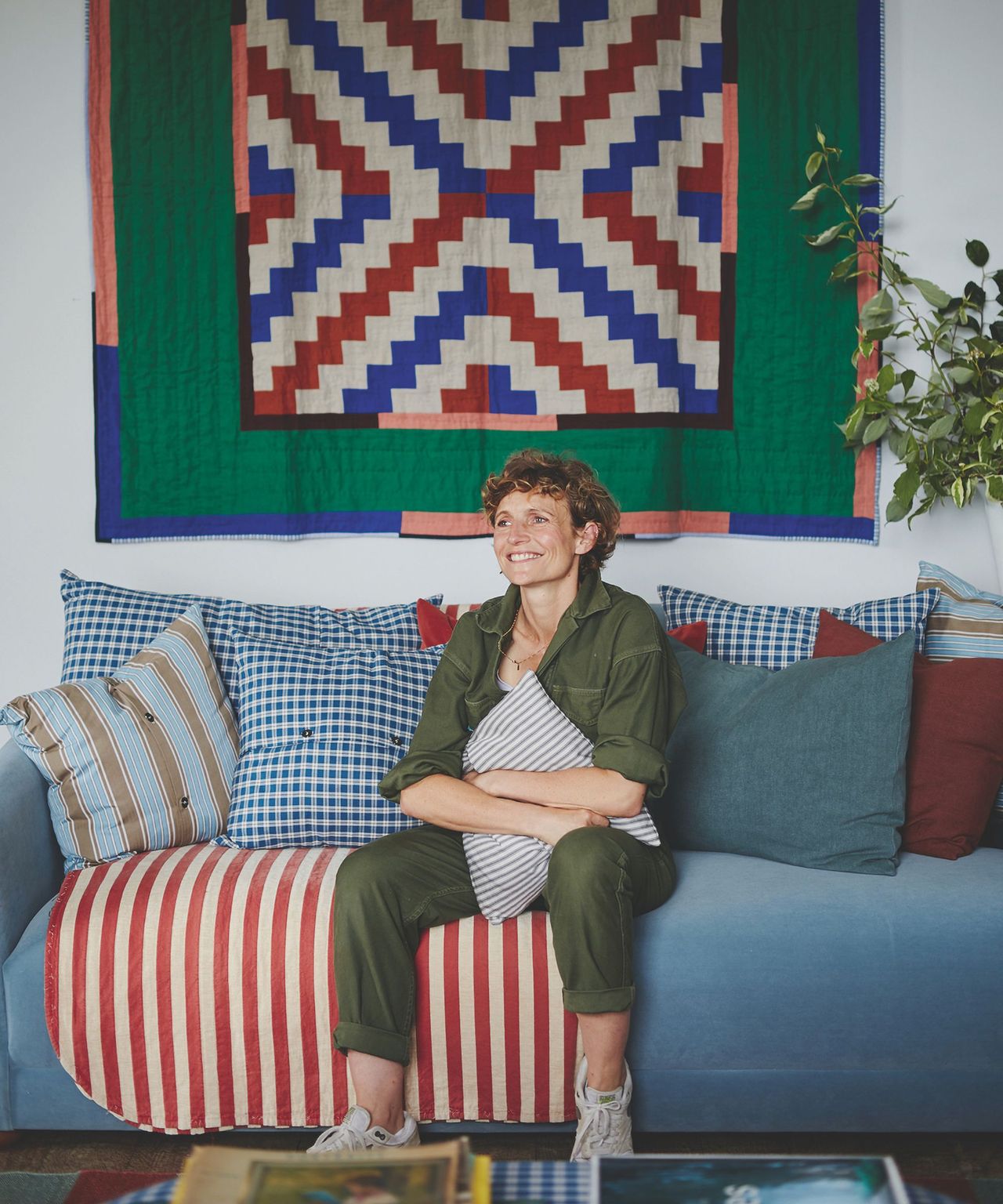 A smiling woman in an olive green boiler suit sits on a blue sofa which is filled with striped scatter cushions. On the wall behind is a patterned wall-hanging