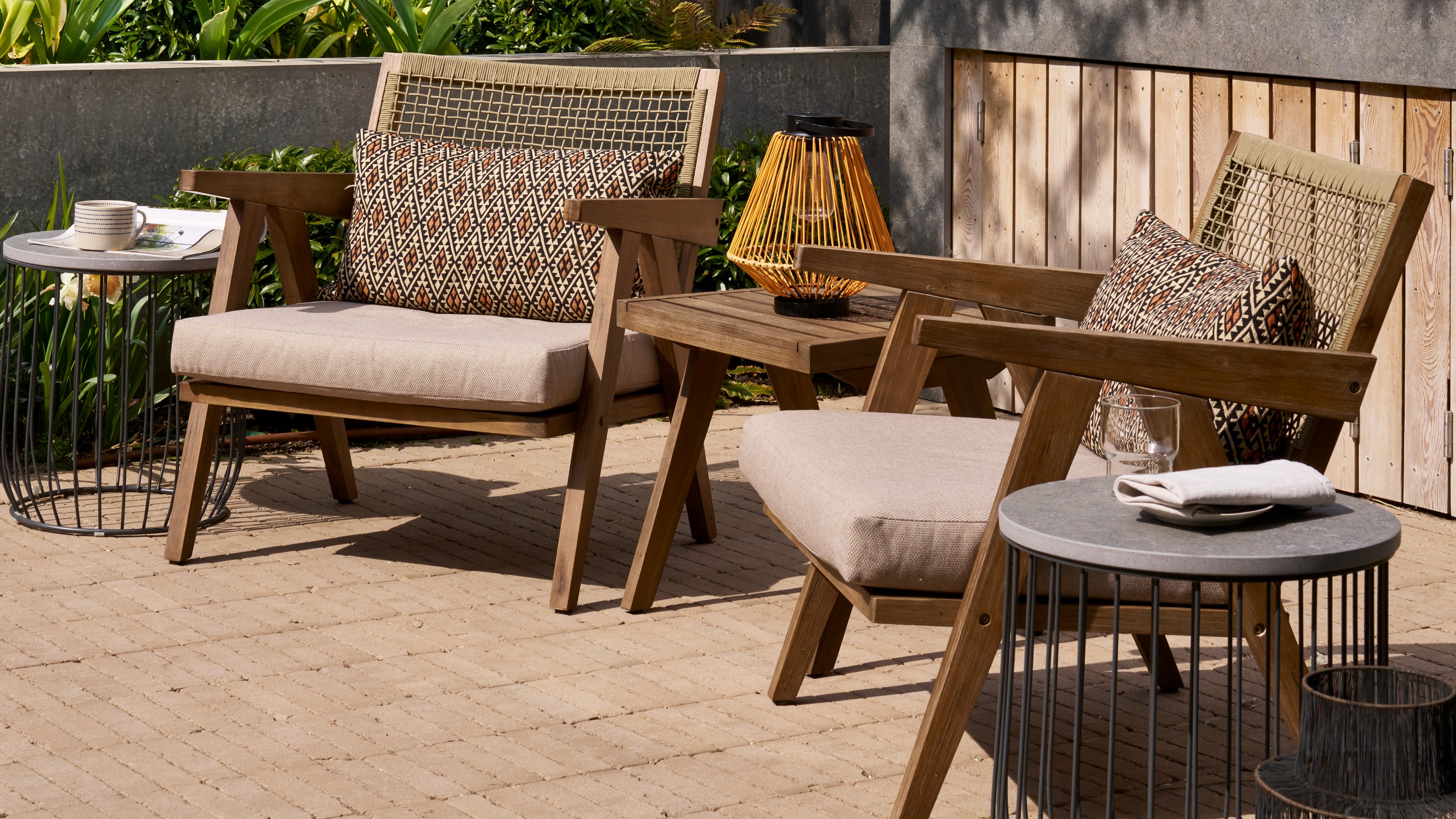 A patio with a set of wooden garden chairs with rattan backs, a matching table and two black metal side tables