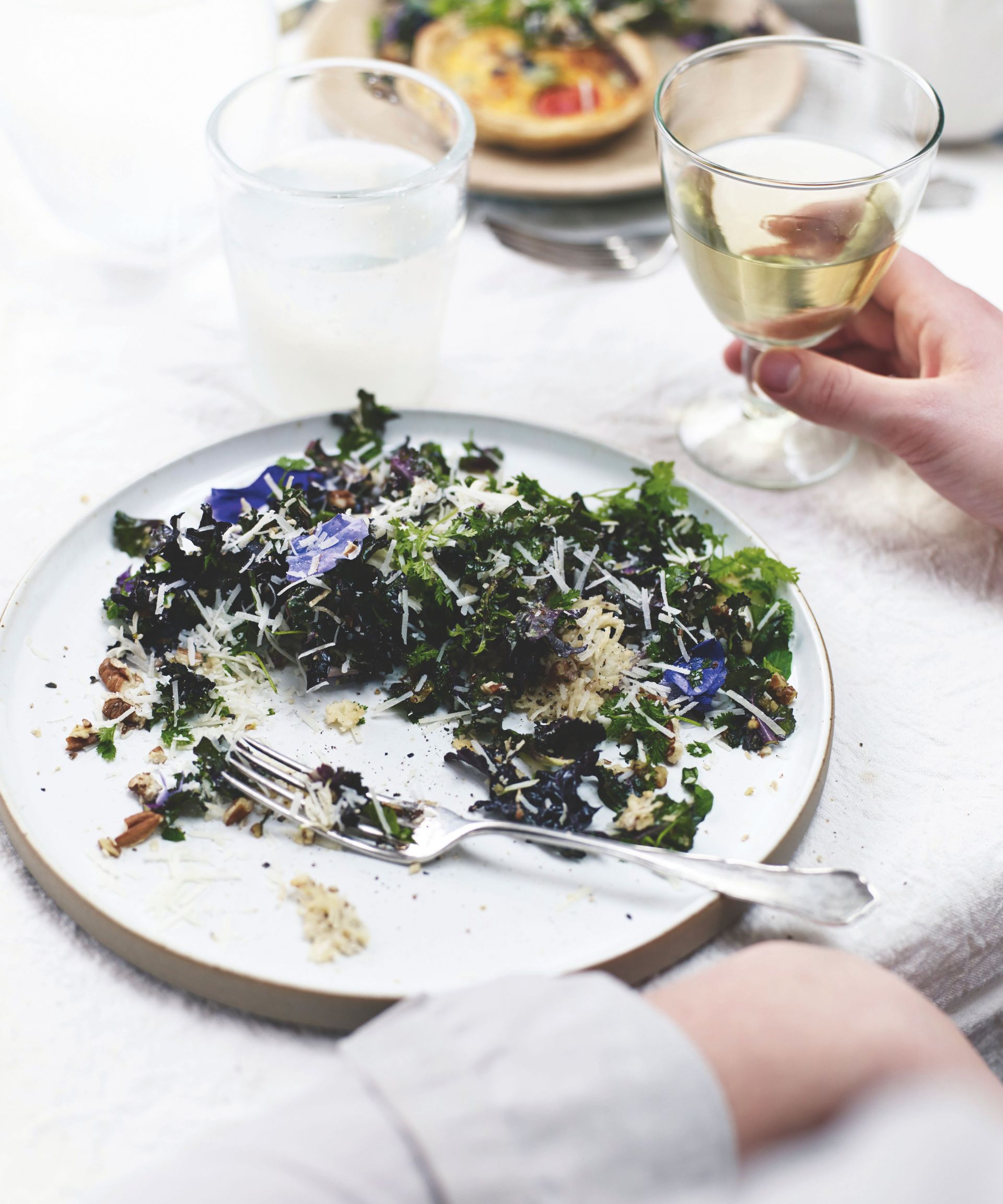 Kale and pecorino crisp salad with edible flowers on a plate on the table with a hand holding a glass of white wine on the table