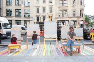 Five easels next to each other with children painting on them and a man sat on a stool