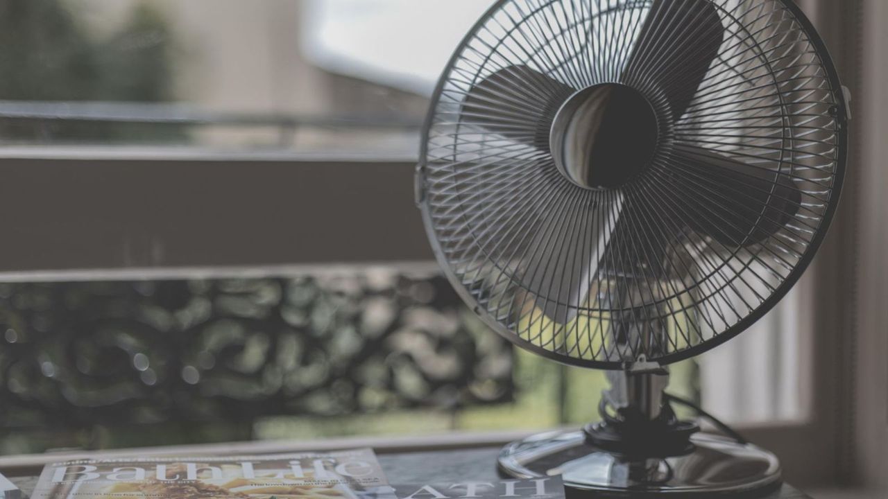 A metal fan next to an open window with a messy stack of magazines beside it