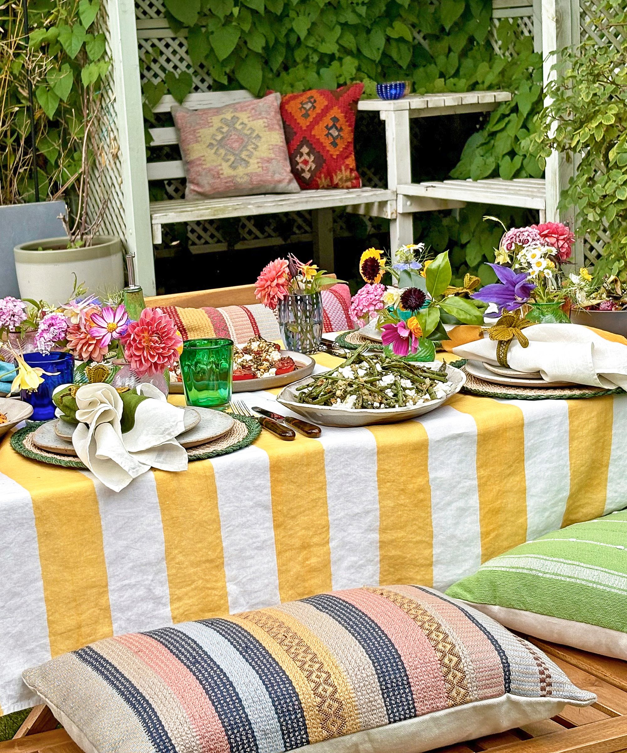 Colorful summer table set up outdoors with flowers, platters of fruit and salads, a yellow and white striped tablecloth and colored glassware with cushions on a bench