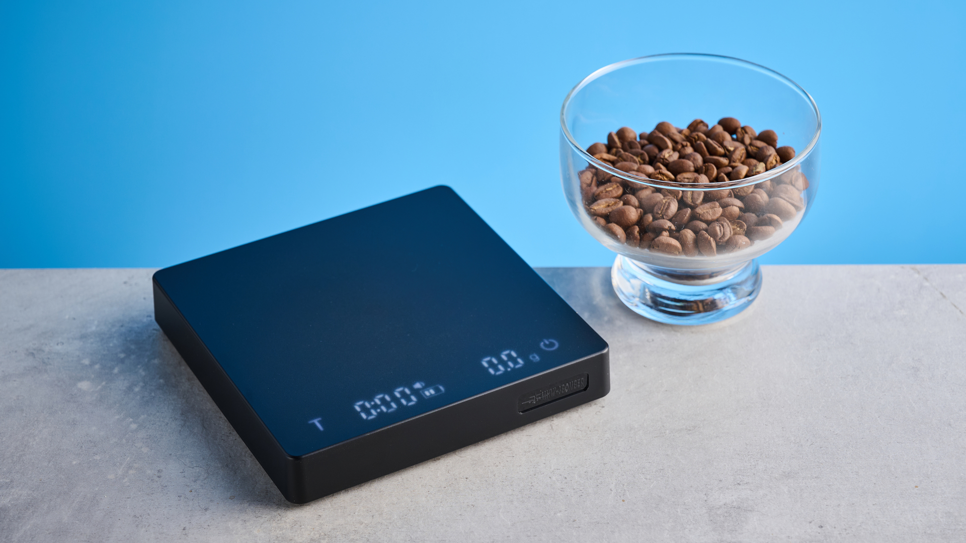 the mhw-3bomber cube coffee scale 2.0 in black photographed against a blue background showing its button controls and dial surface