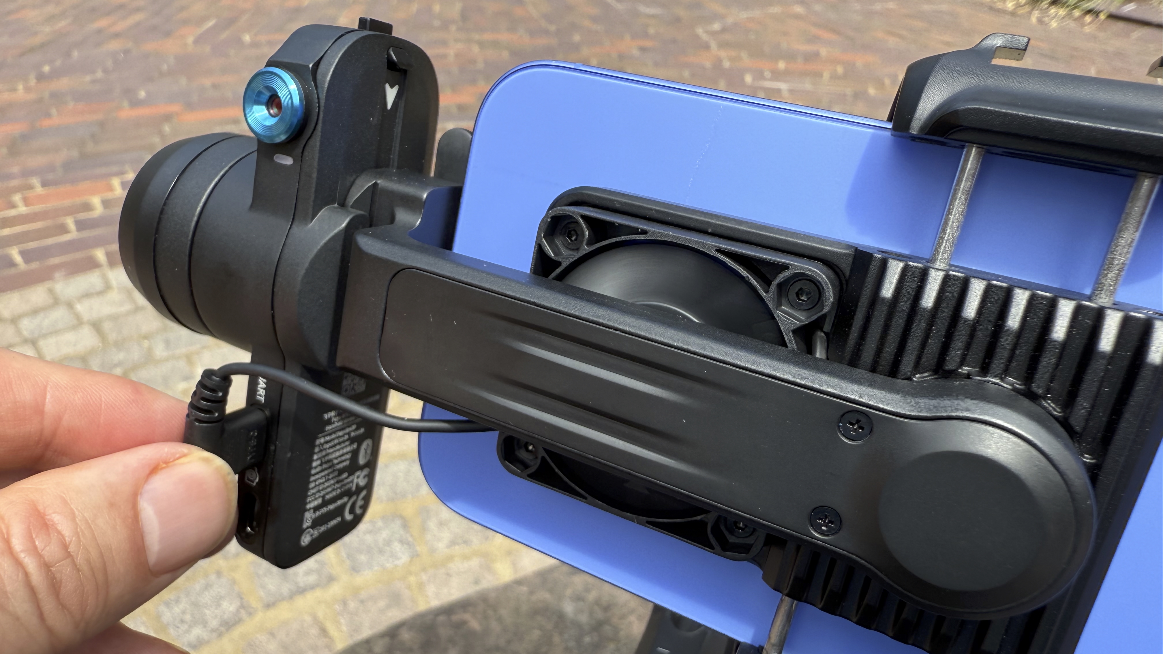 A hand plugs a USB-C cable into a socket on the gimbal to power the smartphone mount’s built-in cooling fan