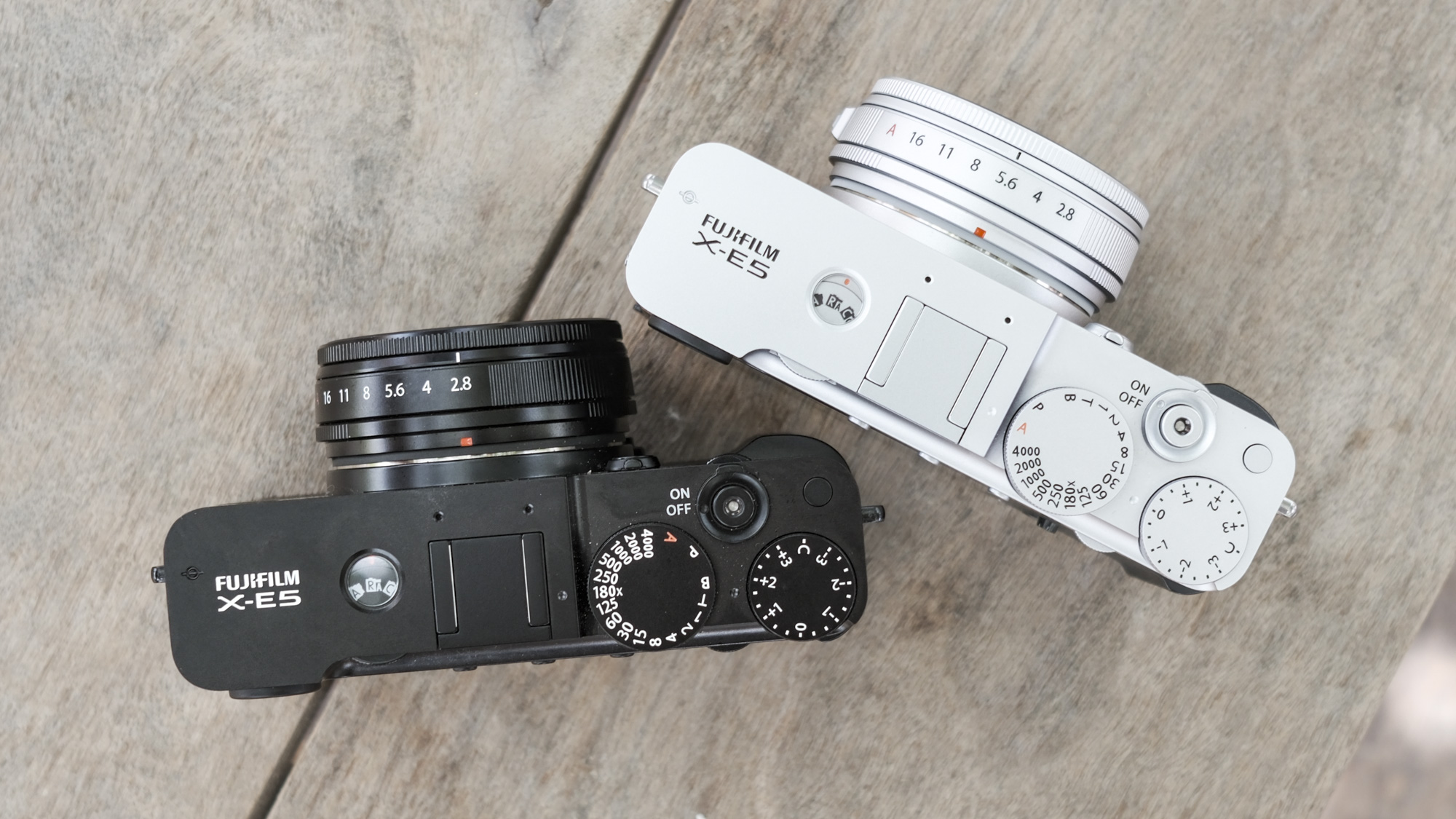 Top down of all black Fujifilm X-E5 alongside silver Fujifilm X-E5, on a wooden bench