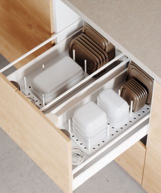 An open light wooden kitchen drawer with white peg organizers in it with clear rectangular containers and brown tub lids neatly stacked within it