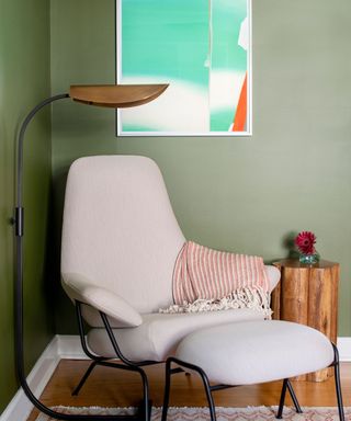 corner of room with olive green walls, neutral armchair with footstool, mint green abstract artwork on the wall and wooden side table