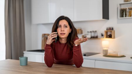 A woman looks frustrated on the phone while sitting at her kitchen island.