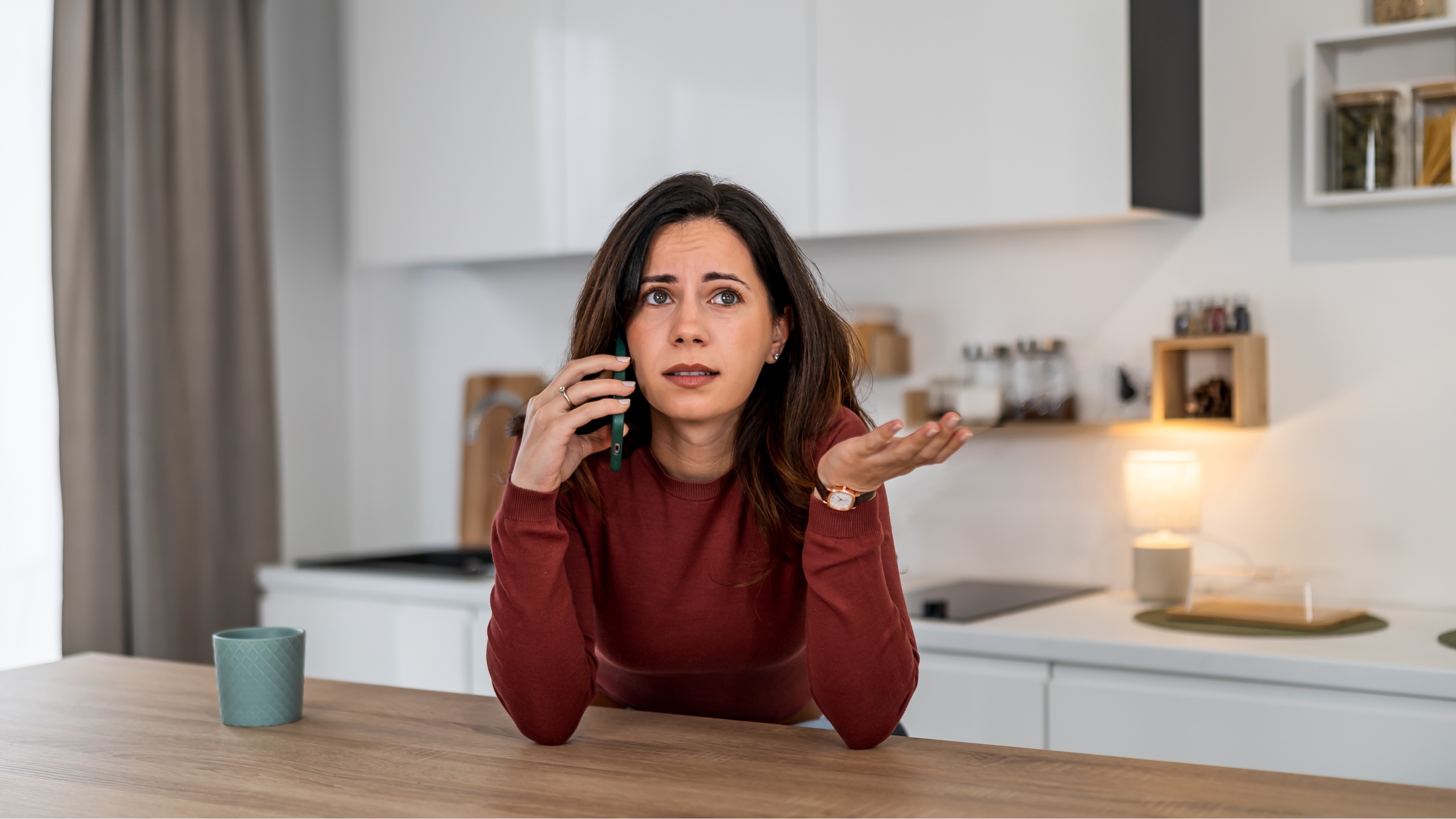 A woman looks frustrated on the phone while sitting at her kitchen island.