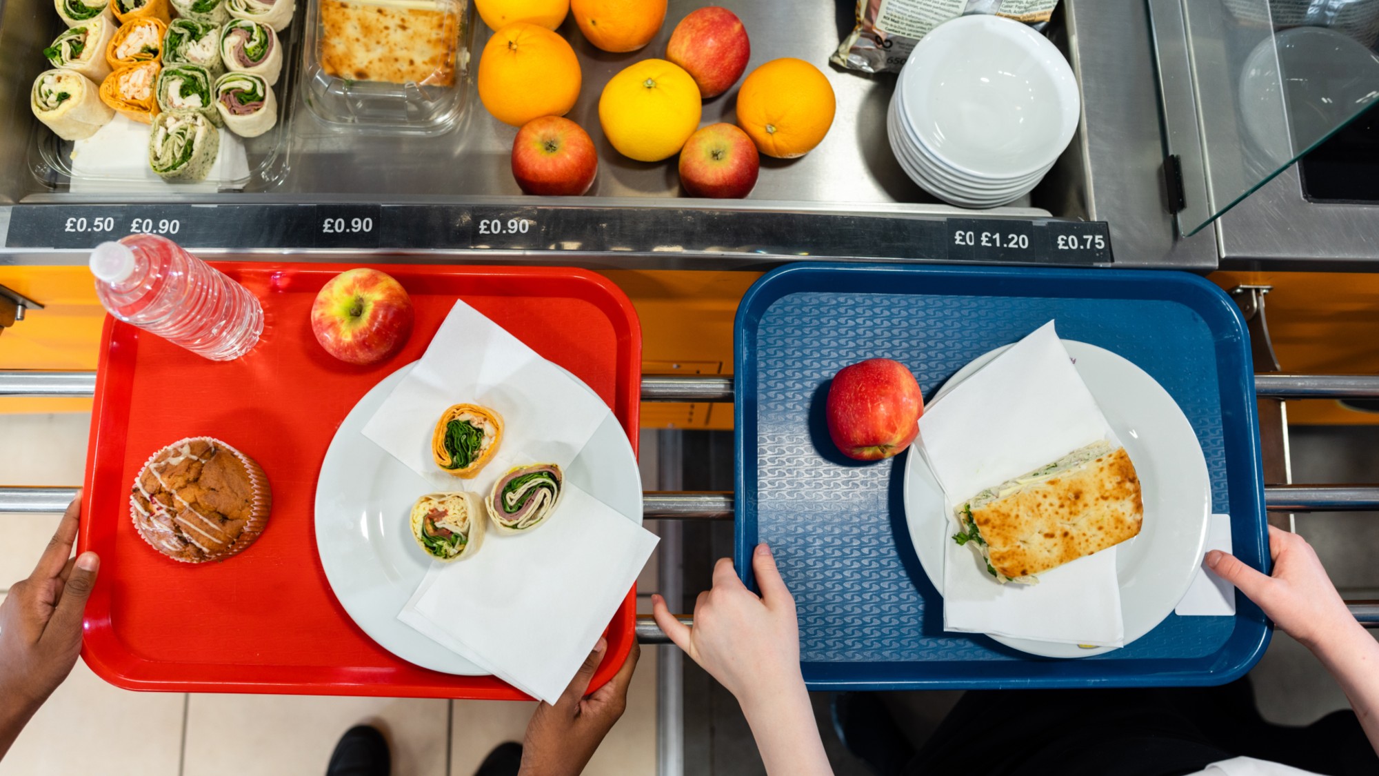 An aerial view of two cafeteria trays containing food items including fruit, wraps and a muffin
