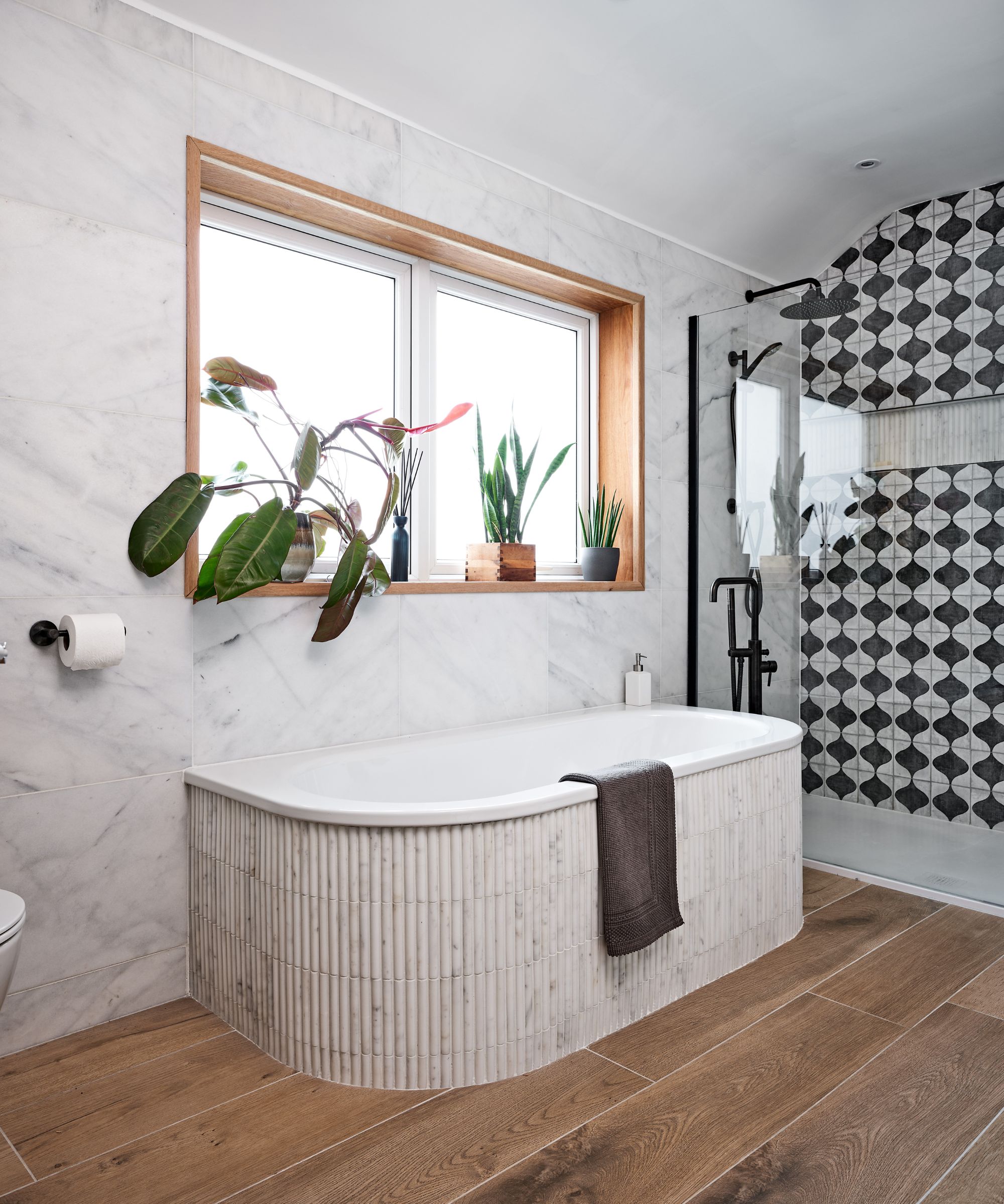 A large neutral bathroom with wooden floors, marble-look tiled walls, fluted tiled on the bath tub and black and white geometric tiles on the shower wall