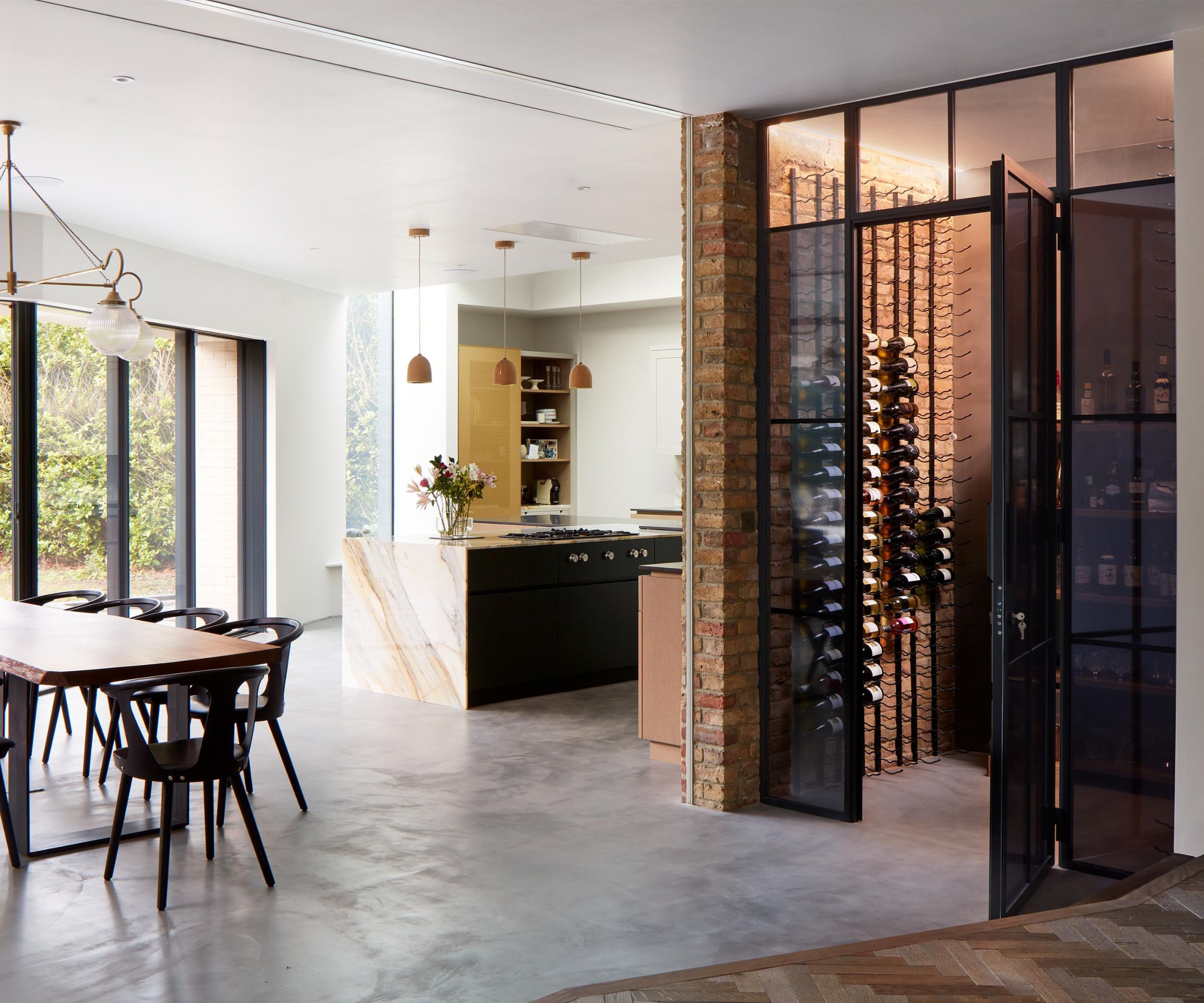 Contemporary open-plan kitchen with a floor-to-ceiling glass wine storage room, exposed brick pillar, marble kitchen island and large dining table with black chairs.