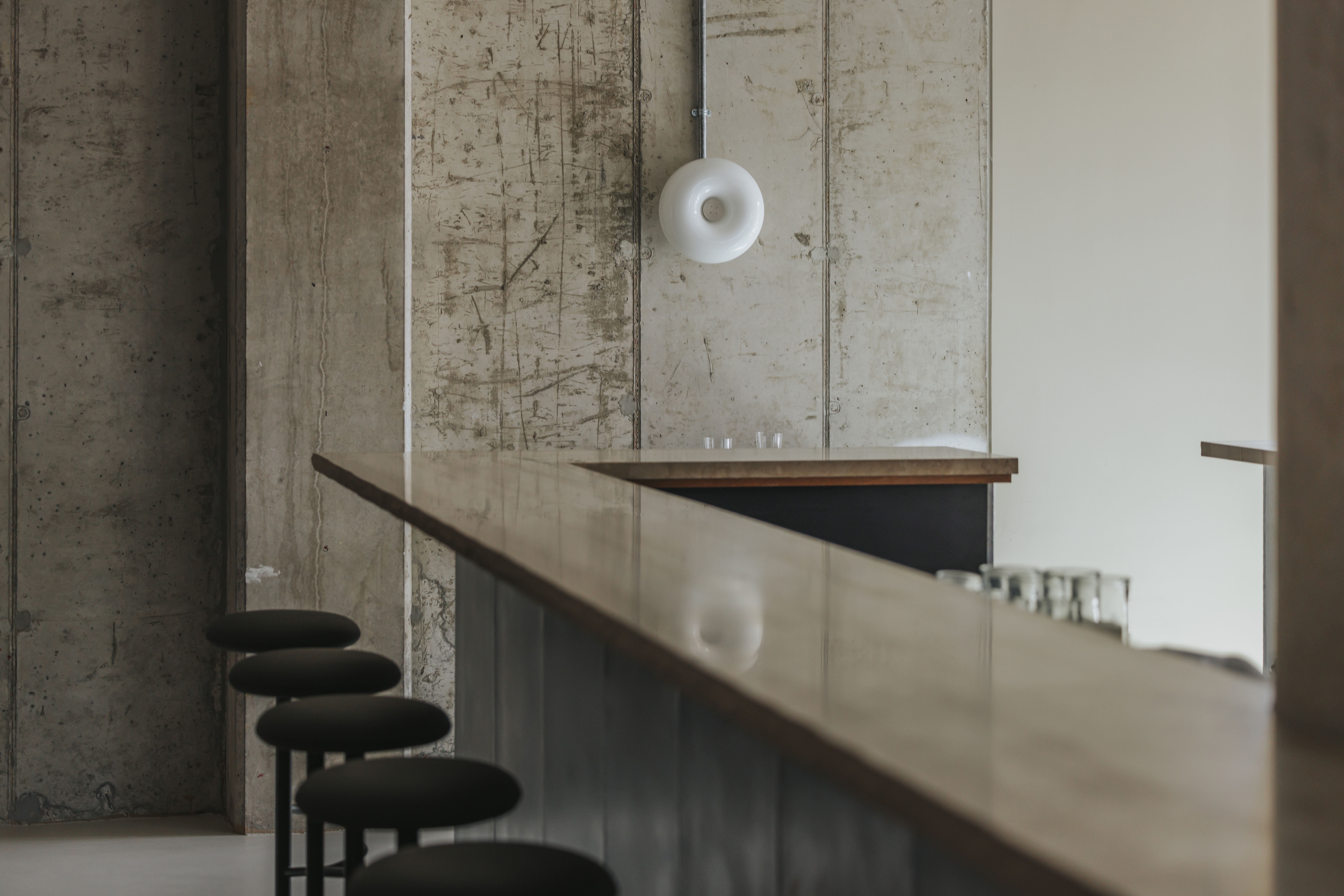 A restaurant bar counter pairs matte metallic surfaces with tall minimalist black stools, a wooden slab, and exposed cement walls adorned with a doughnut-shaped lamp.