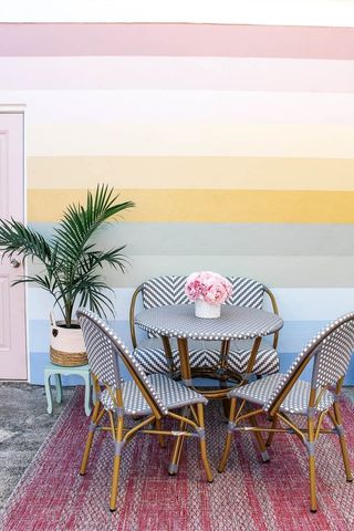 A pink, yellow, and blue striped mural wall with a white and blue rattan woven table and dining chairs and a pink rug underneath