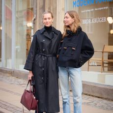 Two women are posing for street style photos in Copenhagen. One woman is wearing a is wearing a black duster trench style coat. The woman is wearing a short cropped coat and jeans. 
