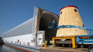 a cone-shaped piece of rocket next to a long, low shelter on top of a barge