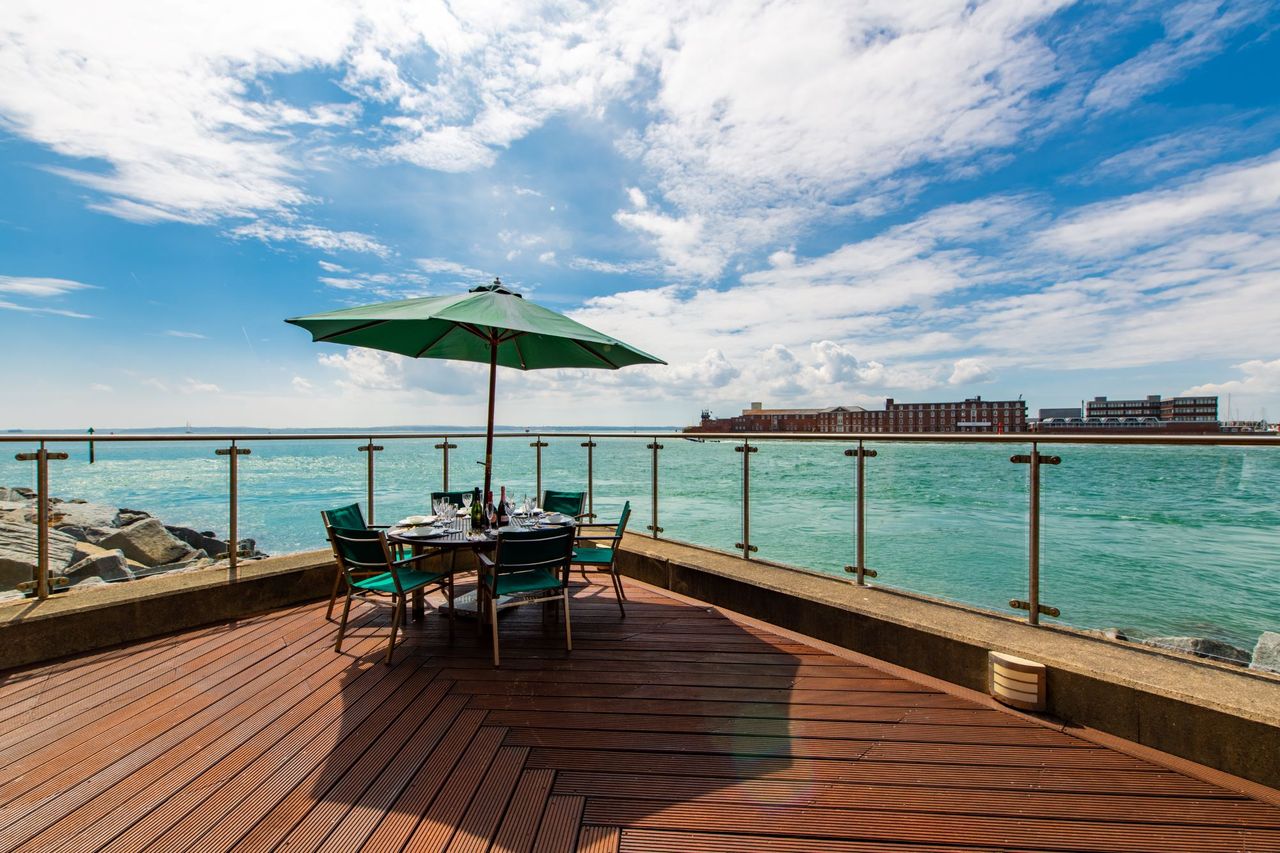 The wooden panelling on the balcony with a wooden table and umbrella looking out onto the sea over a glass panelled boundary