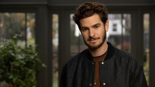 Andrew Garfield, sporting facial hair and wearing a dark grey jacket over a brown t-shirt, looks towards the camera in a promotional image for Who Do You Think You Are Season 22.