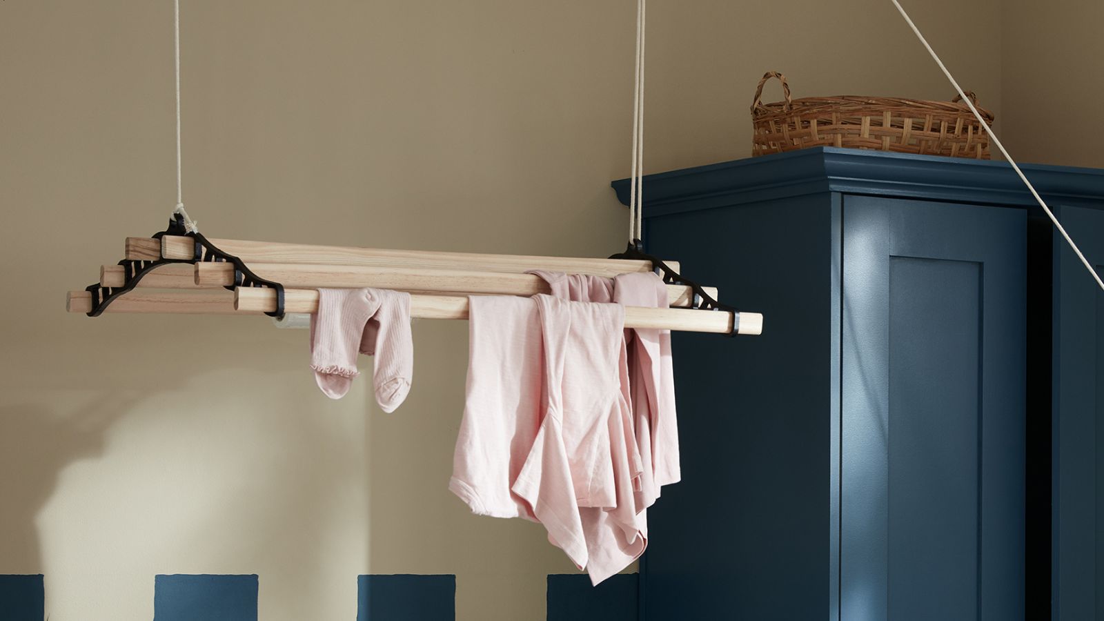 A pale ash wood ceiling-hung pulley airer with black cast iron fittings, draped with pink baby clothes and socks, suspended near a dark teal shaker cabinet.