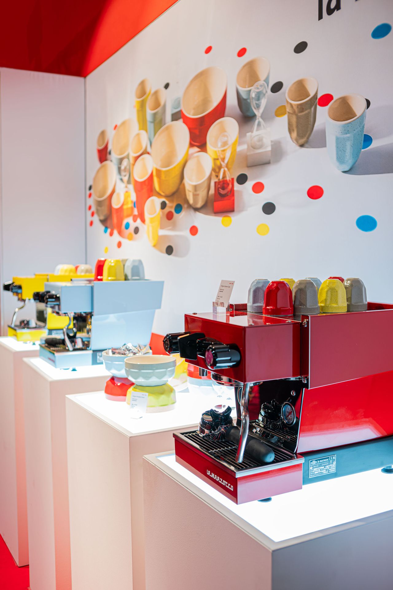 Image of a red, baby blue, and yellow coffee machine each on separate white stand in a room with a poster of matching La Marzocco coffee products behind it.