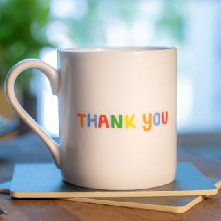 a white mug with the words 'thank you' in rainbow colours written on it, sitting on a wooden kitchen surface