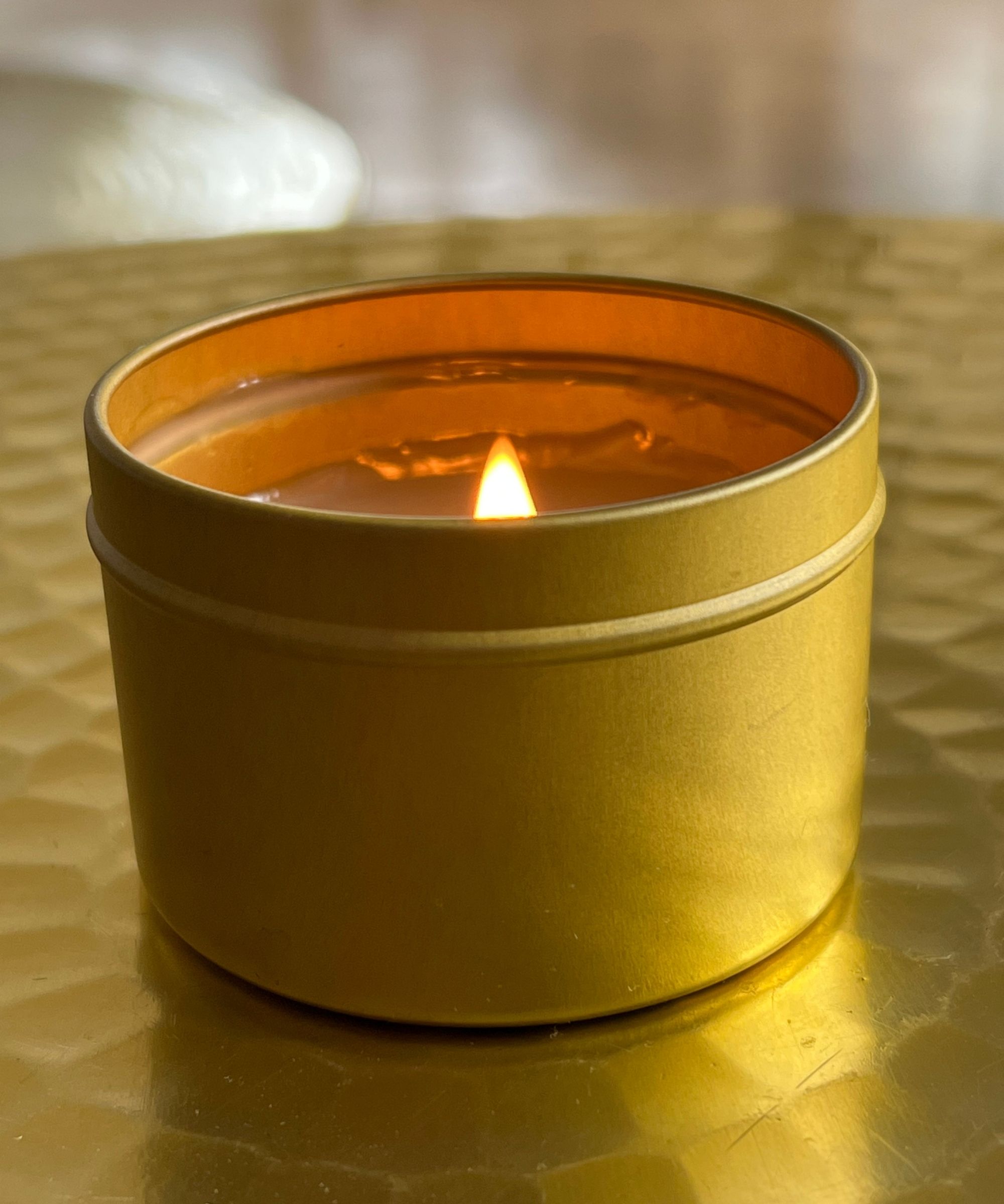 A lit candle in a small gold tin, pictured on a circular hammered gold table, with a bright orange flame.