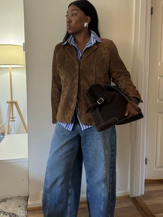 Fashion influencer Nnenna wearing wide-leg jeans, silver statement earrings, a striped button-down shirt, and a brown suede jacket and carrying a suede bag.