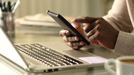 A person holding a phone near a laptop, representing an article about how to set up a Wi-Fi hotspot on Android