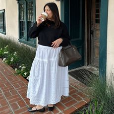 Marina wears white skirt, black sweater, and black flats with a brown handbag while drinking coffee outside of a shop