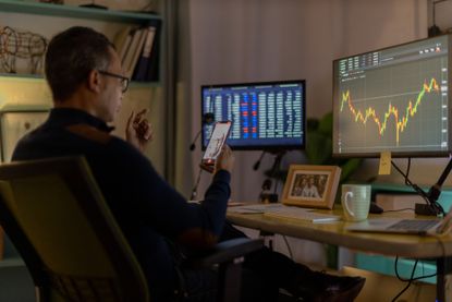 A man holds a phone while sitting at a desk with monitors open to stock performance screens.