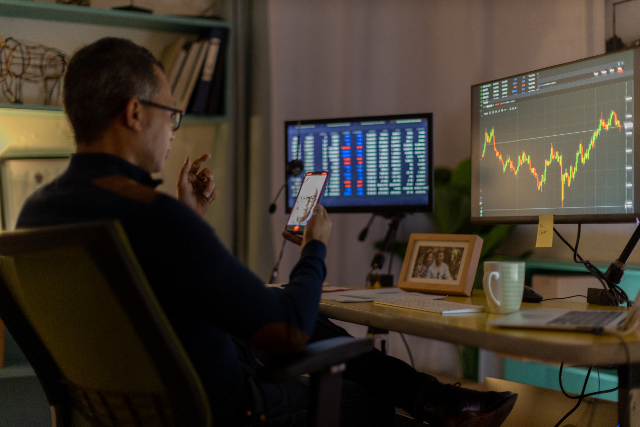 A man holds a phone while sitting at a desk with monitors open to stock performance screens.