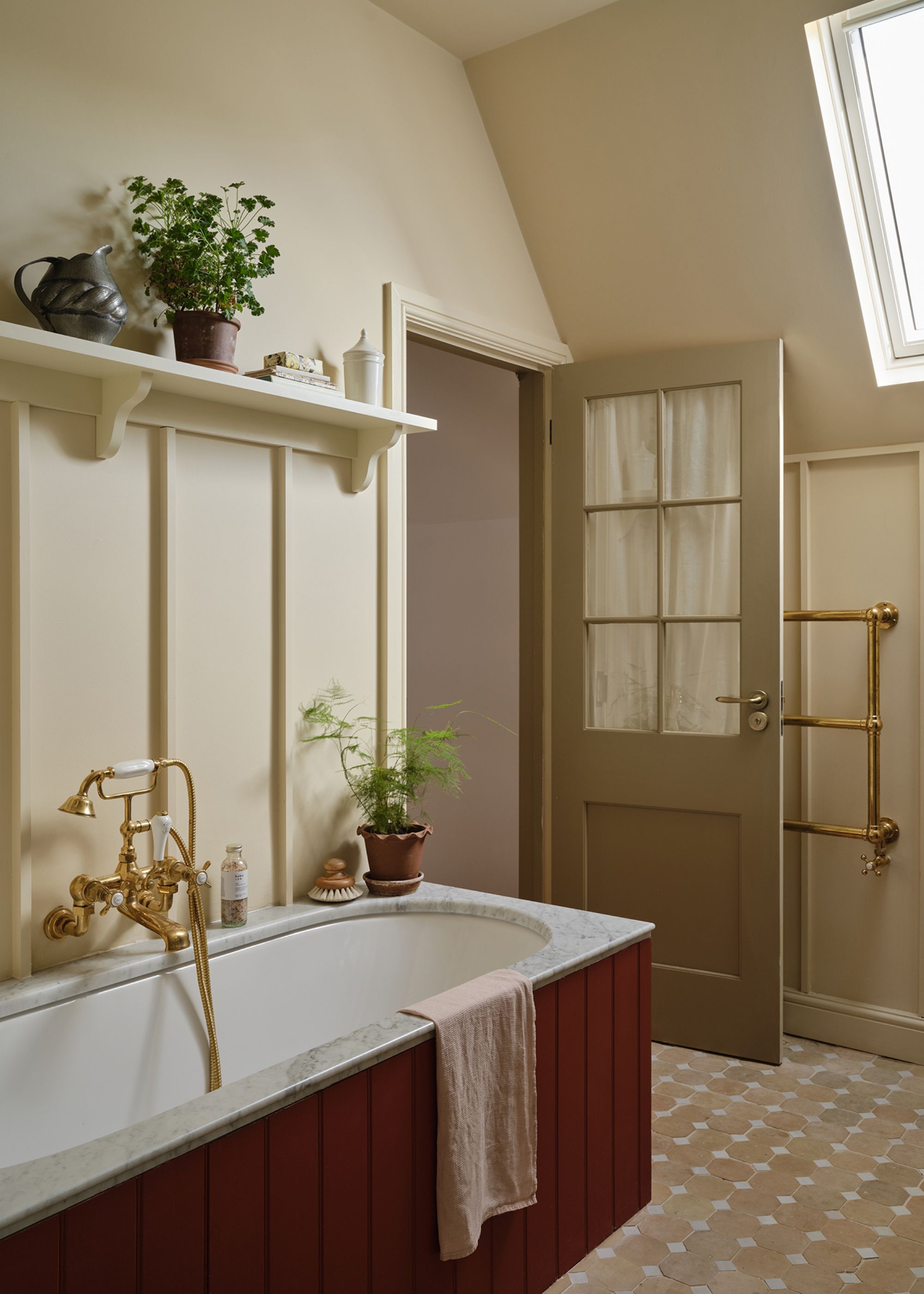 A bathroom with neutral walls and a sloped ceiling with a skylight. Wall panelling with a shelf with decorative objects, and a bathtub with dark red panelled sides and a marble top with brass fixtures