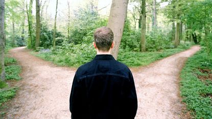 A man stands before a fork in a path in the woods.