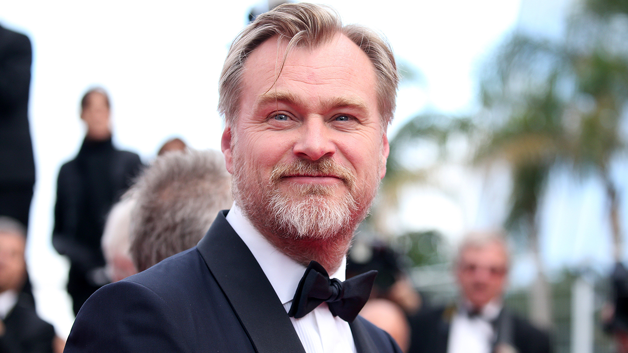 Christopher Nolan smiling in a tuxedo at the Cannes Film Festival, with his hair blowing in the wind.