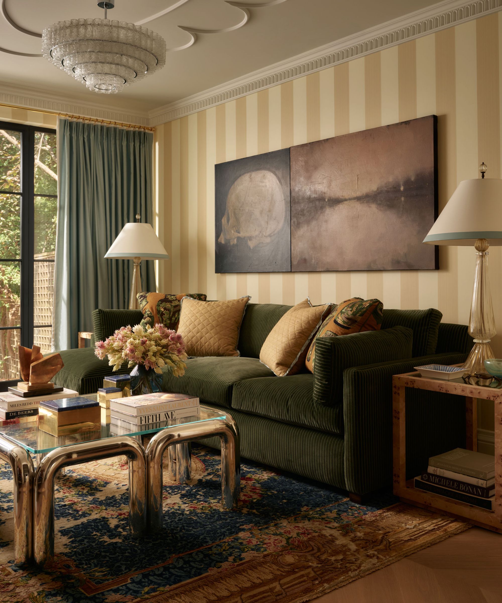a large living room with yellow striped wallpaper, a large green couch, vintage rug, stainless steel coffee table and oversized art and lamps